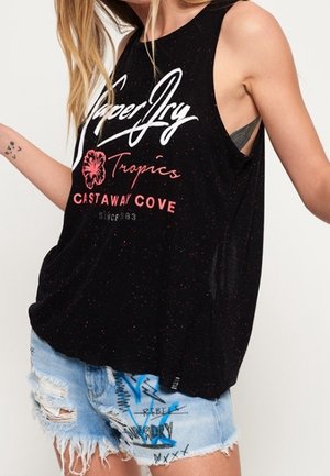 Femme portant un haut noir sans manches avec le texte "Superdry Tropics Castaway Cove" et un short en jean bleu clair déchiré avec des motifs style graffiti.