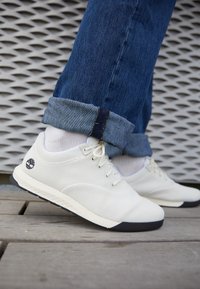 Vita mesh-sneakers med svart sula, med runda snören och en liten logotyp på sidan. Jeans med uppvikta fållar synliga ovanför skon.