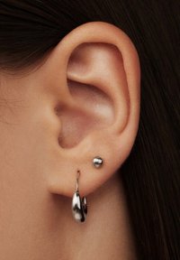 Silberne Ohrringe, die an einem Ohr präsentiert werden: ein kleiner, kugelförmiger Stecker und ein geschwungenes Tropfendesign, beide mit einer reflektierenden, polierten Oberfläche.