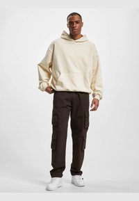 Sudadera crema con un bolsillo frontal, combinada con pantalones cargo marrones que tienen múltiples bolsillos laterales. Zapatillas blancas completan el conjunto.