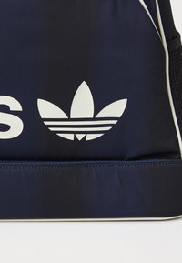 Marineblå sportsbag med teksturert overflate, med et fremtredende hvitt Adidas-logo og kontrasterende kremfarge langs glidelåsen.