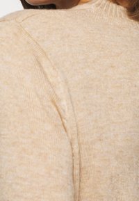 Pull beige en maille avec une finition texturée, doté d'un col côtelé et de légères variations de couleur sur tout le tissu.