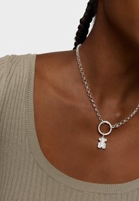 Collier en chaîne argentée avec un lien rond et un pendentif en forme d'ours texturé, porté sur une peau nue près d'un haut côtelé de couleur marron clair.