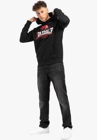 Sudadera con capucha negra con un logotipo gráfico en rojo y blanco de "Lonsdale London", combinada con jeans oscuros y zapatillas deportivas blancas.