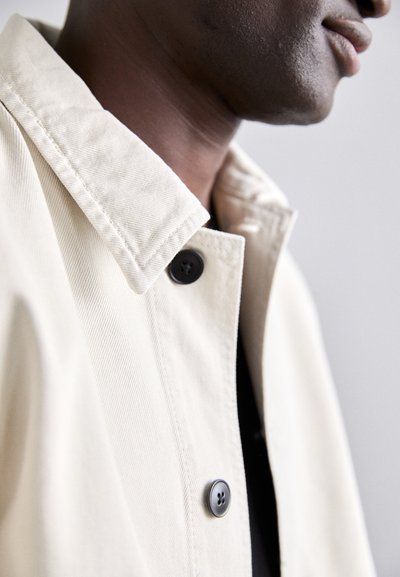 Beige denim jacket with a structured collar, featuring a black button closure and a textured fabric, partially visible in profile.