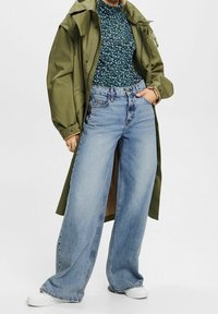 Jeans in denim blu chiaro a gamba larga abbinati a un top floreale verde scuro dalla trama textured e a un giubbotto impermeabile oversize color oliva. Sneakers bianche.