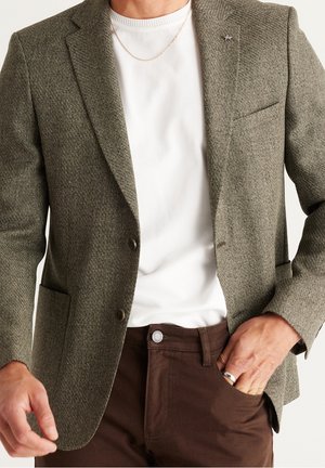 Homme portant un blazer olive texturé sur une chemise blanche, un pantalon marron, un collier chaîne argenté et une bague à la main droite.