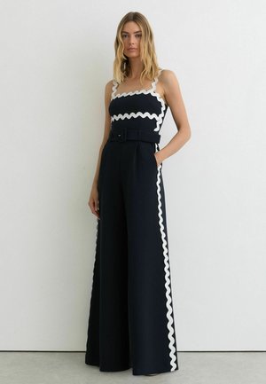 Kvinde iført en sort ærmeløs jumpsuit med vide ben og hvid bølget kant langs kanterne, stående mod en ensfarvet hvid væg.