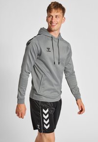 Grijze hoodie met lange mouwen, een voorklep en zwarte accenten. Gecombineerd met zwarte shorts met witte chevronstrepen aan de zijkant.