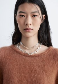 Silberne Kettenhalsketten, die über einem braunen, flauschigen Pullover geschichtet sind, mit glatter Textur und runden Gliedern, die am Hals eine strukturierte Optik verleihen.