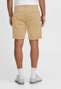 Pantaloni scurți kaki cu manșete răsucite, din bumbac, cu două buzunare la spate cu închidere cu nasture. Purtați cu adidași gri și șosete albe.