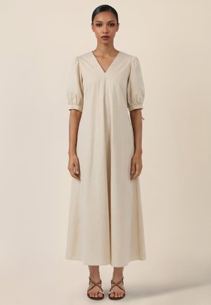 Frau steht und trägt ein langes beiges Kleid mit aufgebauschten, ellbogenlangen Ärmeln und V-Ausschnitt, kombiniert mit braunen, geschnürten Sandalen.