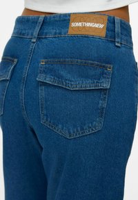 Los jeans de mezclilla azul oscuro presentan un parche cuadrado de cuero etiquetado como "SOMETHINGNEW" en la cintura trasera, con dos bolsillos traseros y costuras amarillas.