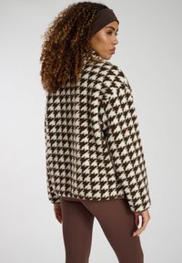 Giacca in pile a motivo houndstooth marrone e bianco con collo alto e vestibilità rilassata, abbinata a leggings marroni. Design texturizzato e accogliente.