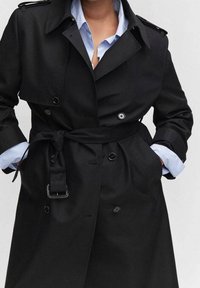 Czarny trencz z paskiem, dwurzędowymi guzikami, szerokimi klapami i gładką fakturą. Częściowo widoczna jest jasnoniebieska koszula pod spodem.