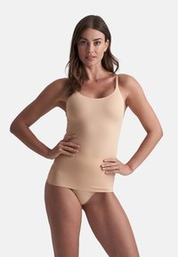 Mujer vestida con un camisón nude sin costuras y bragas tipo thong, posando con las manos en las caderas frente a un fondo liso claro.