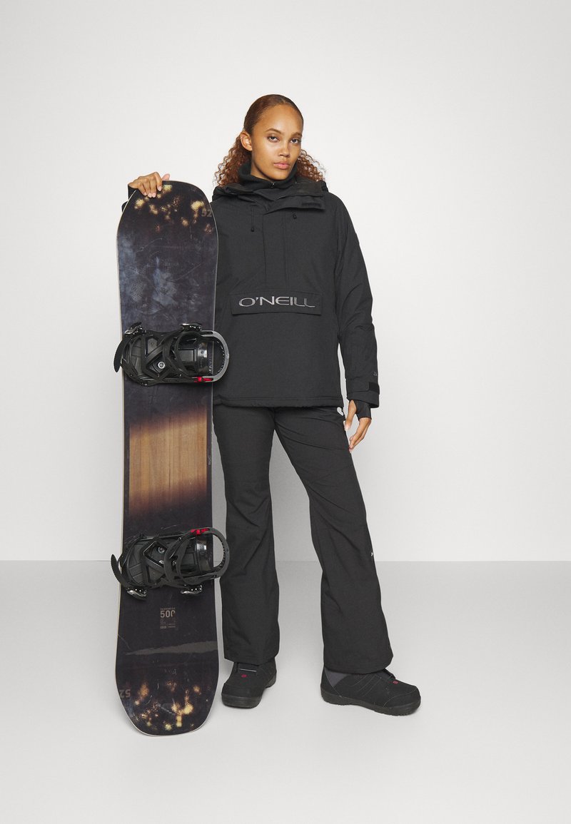 Schwarze Snowboardjacke mit einem Logo auf der Vorderseite, schwarze Hose und schwarze Stiefel. Hält ein Snowboard mit dunklen und hellen Holzmustern und schwarzen Bindungen.