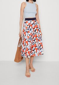 Top cropped azzurro chiaro, gonna plissettata a fantasia in blu navy, bianco, arancione e azzurro chiaro, abbinati a sandali intrecciati beige e una borsa a tracolla marrone.