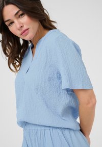 Light blue, textured short-sleeve top with a v-neck design and relaxed fit. The fabric appears crinkled with a subtle softness.
