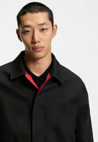 Musta takki, jossa on punainen sisävuori, kauluksellinen pääntie ja napillinen sulkus. Kangas näyttää sileältä ja kestävältä.