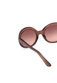 Lunettes de soleil rondes à monture brune translucide avec verres dégradés et logo "Guess" sur la branche, inclinées sur fond blanc.