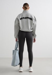Grå oversized genser med svart tekst "AIM'N APPAREL" på baksiden, kombinert med svarte ribbestrikkede leggings og en lyseblå handlenett.