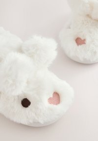 Paire de chaussons blancs et duveteux en forme de lapin avec des yeux brodés et des nez roses en forme de cœur sur une surface claire.