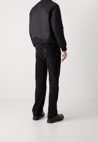 Schwarze Bomberjacke mit gerippten Bündchen, kombiniert mit schwarzen Jeans. Auffälliges rotes Markenetikett an der Rückentasche. Die Schuhe sind schwarz mit einer glatten Oberfläche.