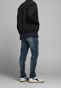 Černá bomber bunda, modré tričko pod ní, prošlé modré džíny a bílé tenisky s černými akcenty. Hladká látka, uvolněný střih.