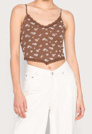 Bruine cropped tanktop met een wit bloemenpatroon, geschulpte kantafwerking en spaghettibandjes, gecombineerd met hoge witte jeans.