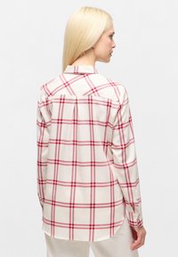 Woman with long blonde hair wearing a cream shirt with red plaid pattern, shown from behind against a plain light background.