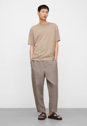 Junger Mann steht mit den Händen in den Taschen, trägt ein beigefarbenes Kurzarmhemd, lockere braune Hose und dunkelbraune Sandalen vor einem einfarbigen hellen Hintergrund.