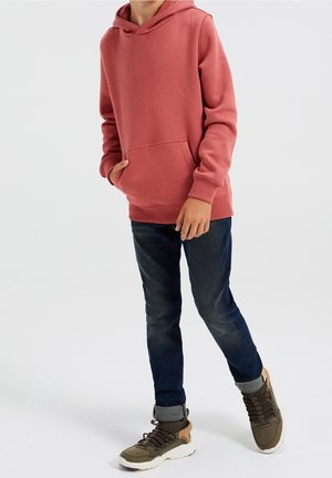 Sweat à capuche rose avec poche frontale, associé à un jean en denim foncé et des baskets vert olive avec des détails texturés et des lacets.