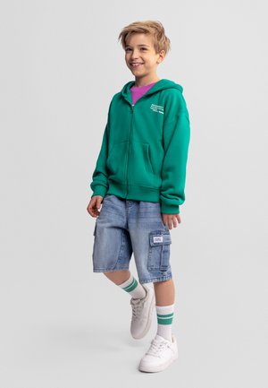 Uśmiechnięty chłopiec w zielonej bluzie na zamek, fioletowej koszulce, dżinsowych krótkich spodniach typu cargo, białych trampkach i białych skarpetkach z zielonymi paskami, stojący na jednej nodze.