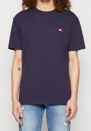 Navyblauw katoenen t-shirt met korte mouwen, ronde halslijn en een klein logo-opdruk op de linkerkant van de borst. Gecombineerd met versleten lichtblauwe jeans.