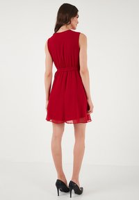 Red sleeveless dress with a fitted waist and flowing skirt. Made of lightweight fabric, featuring a solid color and a smooth texture.