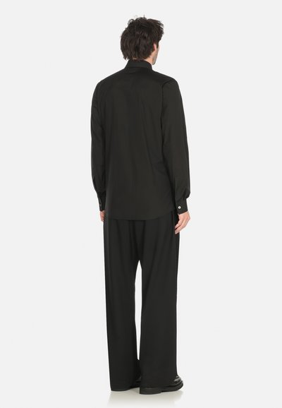 Uomo in piedi di spalle, che indossa una maglietta nera a maniche lunghe e pantaloni neri a gamba ampia, su uno sfondo bianco semplice.