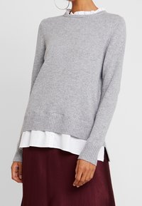 Grauer Strickpullover mit Rippabschluss und -ärmeln, der einen runden Halsausschnitt und seitliche Schlitze aufweist. Getragen über einem schichtweisen weißen Hemd und einem bordeauxroten Rock.