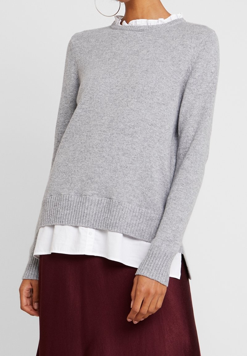Grauer Strickpullover mit Rippabschluss und -ärmeln, der einen runden Halsausschnitt und seitliche Schlitze aufweist. Getragen über einem schichtweisen weißen Hemd und einem bordeauxroten Rock.