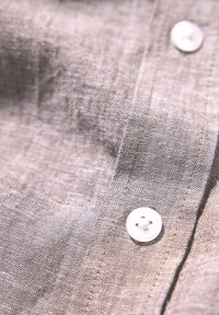 Vaaleanharmaa pellava, jossa on teksturoitu pinta, valkoisella napilla ja ommellulla sauman yksityiskohdalla. Lähikuva materiaalista.