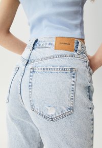 Persona vistiendo una camiseta de tirantes de canalé azul claro y unos vaqueros de tiro alto de lavado claro con un pequeño desgarro y una etiqueta marrón de Pull&Bear en la cinturilla.