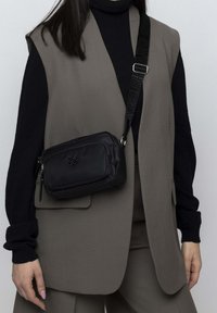 Schwarze Umhängetasche aus Nylon; verfügt über ein Reißverschlussfach, ein Logo und einen verstellbaren Riemen mit einem strukturierten Muster.