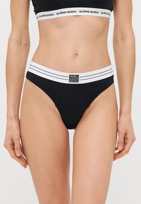 Culotte de bikini noire avec une ceinture élastique rayée blanche. Présente une étiquette logo à l'avant. Fabriquée en matériau lisse.