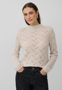 Femme aux longs cheveux bruns portant des boucles d'oreilles en or, un pull en tricot beige à motifs et un jean foncé, les mains dans les poches, sur fond gris.