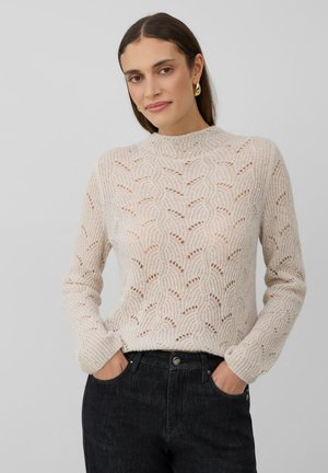 Vrouw met lang bruin haar die gouden ringoorbellen draagt, een beige gebreide trui met patroon en donkere spijkerbroek, met haar handen in de zakken, tegen een grijze achtergrond.