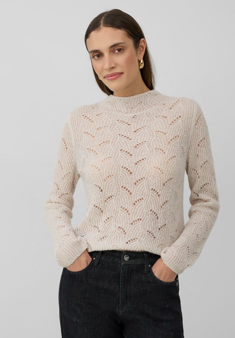 Femme aux longs cheveux bruns portant des boucles d'oreilles en or, un pull en tricot beige à motifs et un jean foncé, les mains dans les poches, sur fond gris.