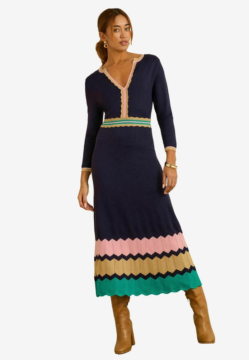 Strikkekjole i marineblå med V-hals, bølgete detaljer og chevronmønstre i pink, guld, grøn og sort ved kanten. Lange ærmer.
