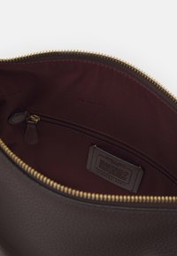 Sac en cuir marron foncé ouvert montrant un intérieur en tissu bordeaux avec une poche zippée et une étiquette de marque cousue à l'intérieur.