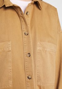 Camicia button-up color beige realizzata in tessuto morbido, con due tasche anteriori, dettagli di cucitura visibili e colletto arrotondato.
