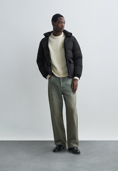 Vyras vilkintis juodą pūkinę striukę, užsidėjęs kreminės spalvos megztinį, laisvas išblukusias žalias džinsus ir juodus batus, stovintis prieš šviesų vienspalvį foną.